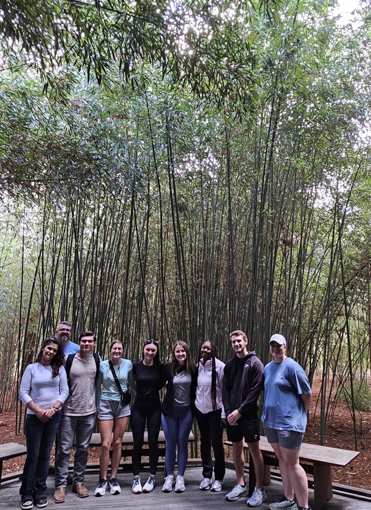 Eight students and an instructor standing in front of a grove of tall thin trees.